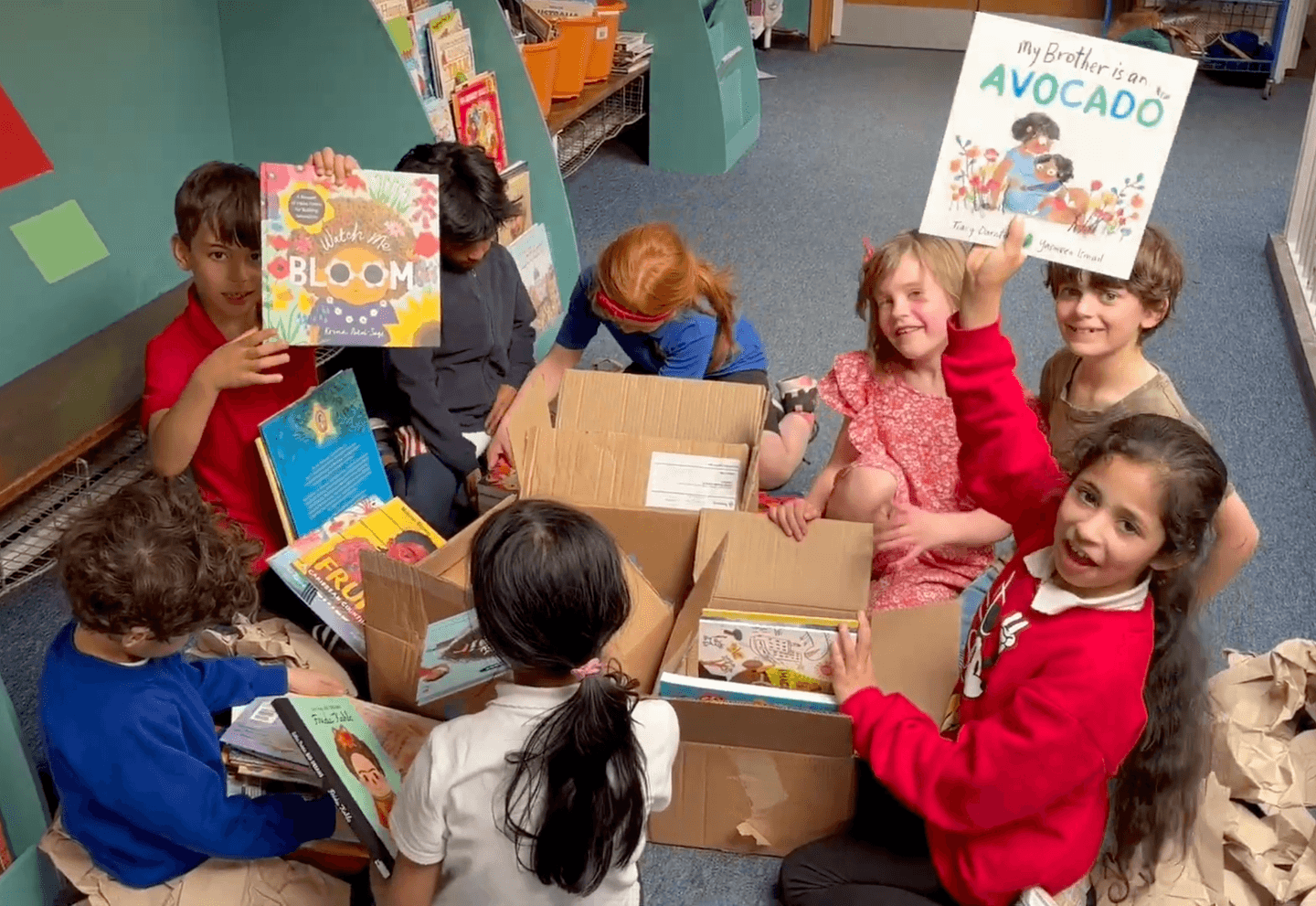 A photo of 8 children in a school library excitedly unboxing their IBC Book Box. 4 children grin at the camera, 2 of whom are waving books in the air. 
