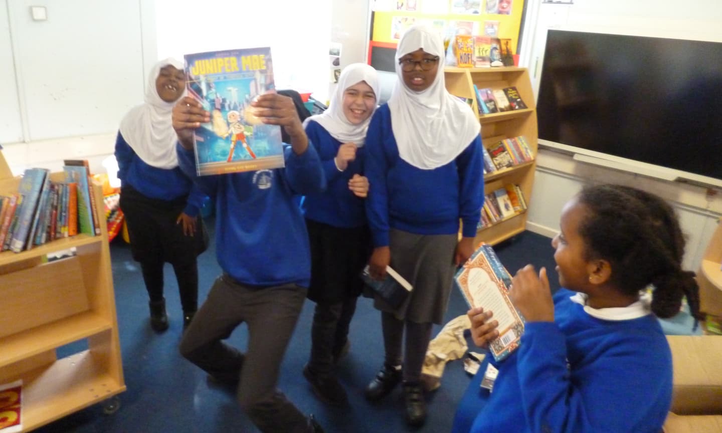 A photo of uniformed children in a school library, who have just been unboxing their IBC Book Box, A young Black boy strikes a pose, brandishing a copy of the graphic novel Juniper Mae (which hides his face). His peers, 3 girls in Hijabs, and a fourth, a girl with brown skin holding a book, laugh happily around him.