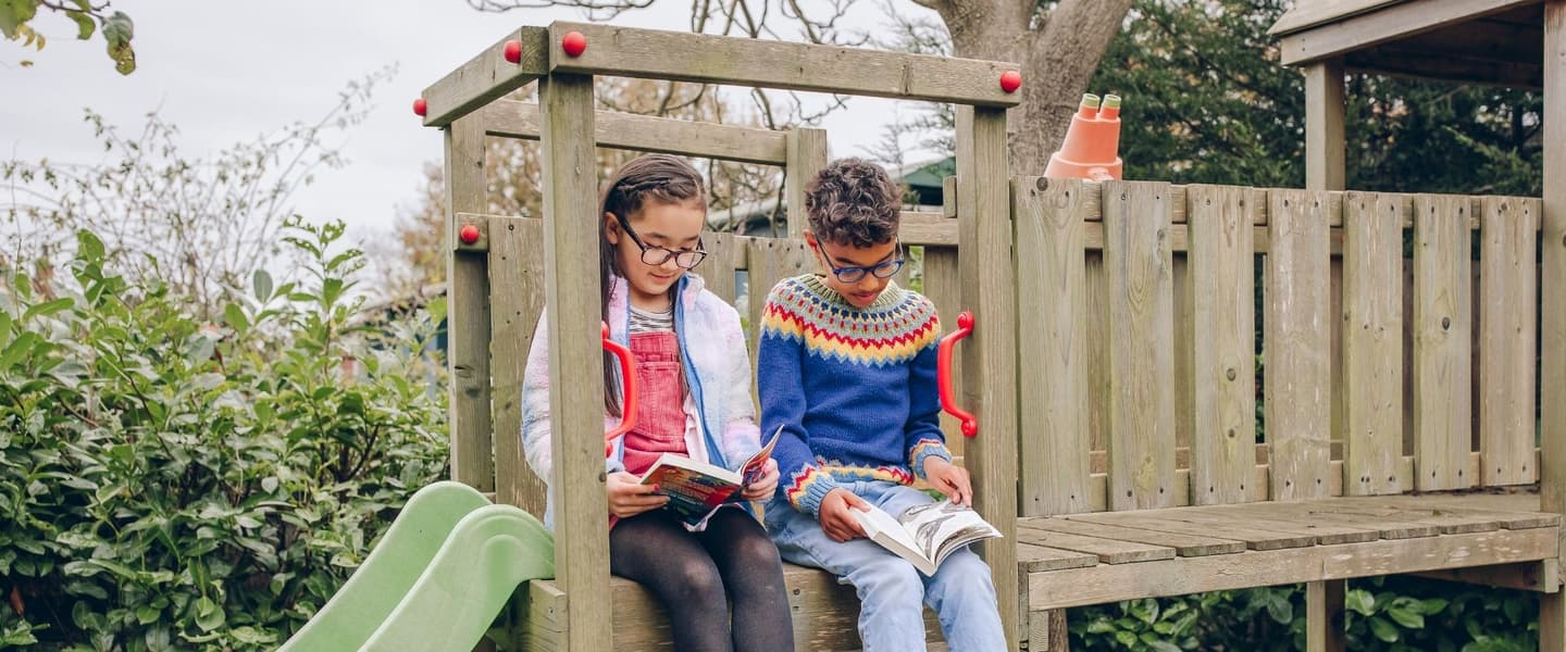 Two children, a mixed white and East Asian girl and a mixed white and Black boy, are sitting atop a climbing frame with a green slide in a garden. They are both smiling and reading children’s novels. 
