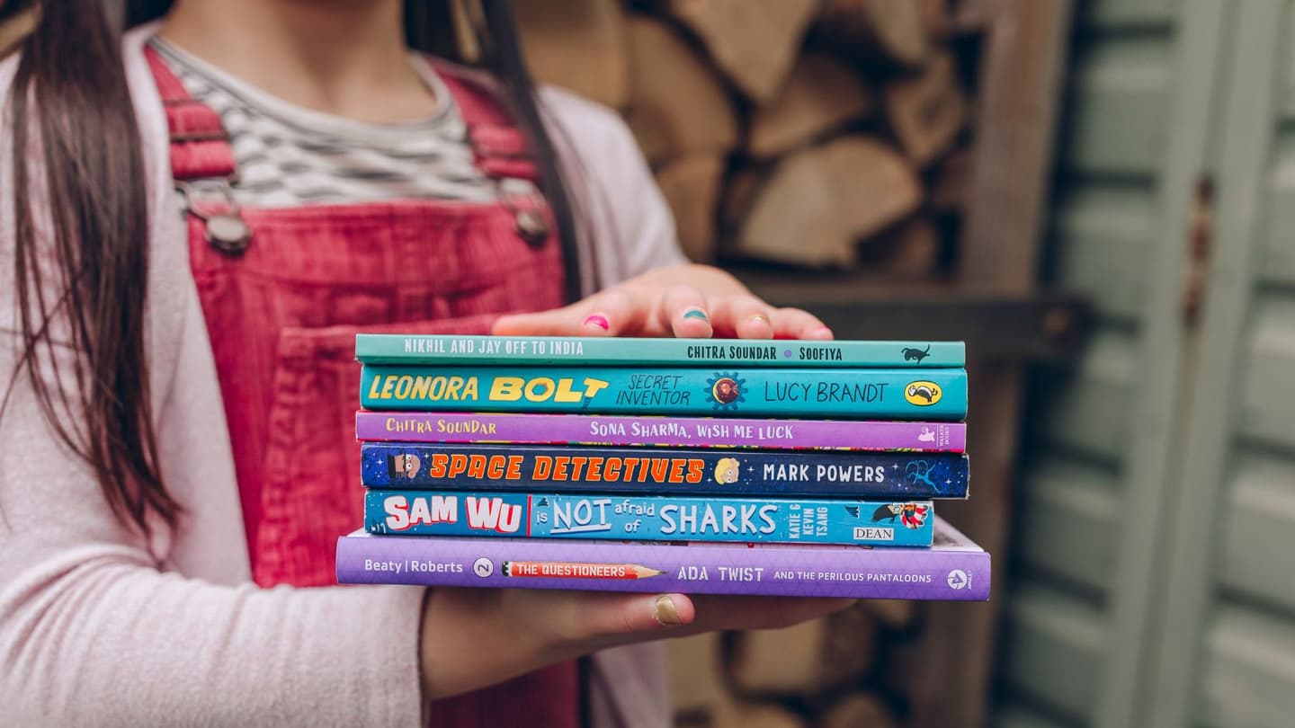 An Asian girl holding a pile of children's books outside by a fence. 