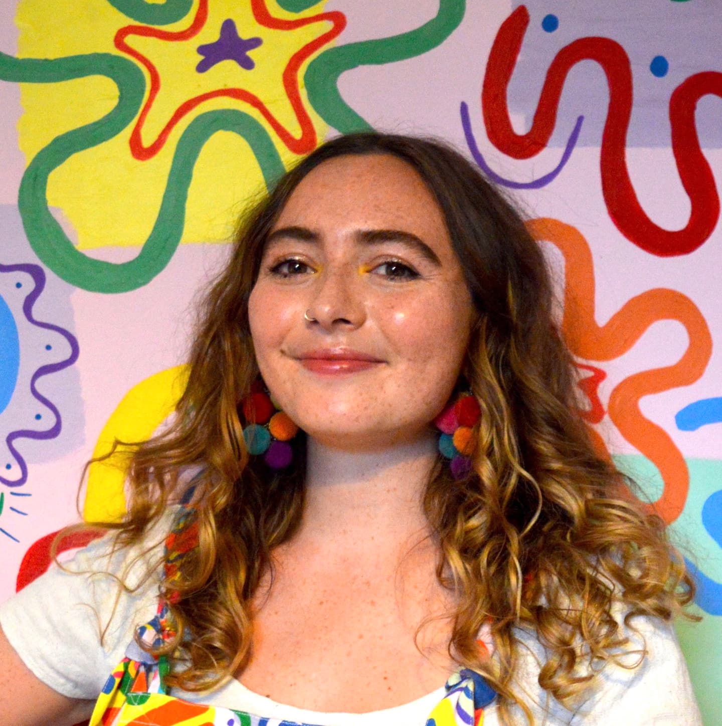 Molly, a woman in her twenties standing in front of a brightly coloured mural (one of her mural painting designs). She wears dungaress to match the mural pattern behind her. She has wavy brown hair with some blonde, wears colourful pom pom earrings and smiles happily.\n
