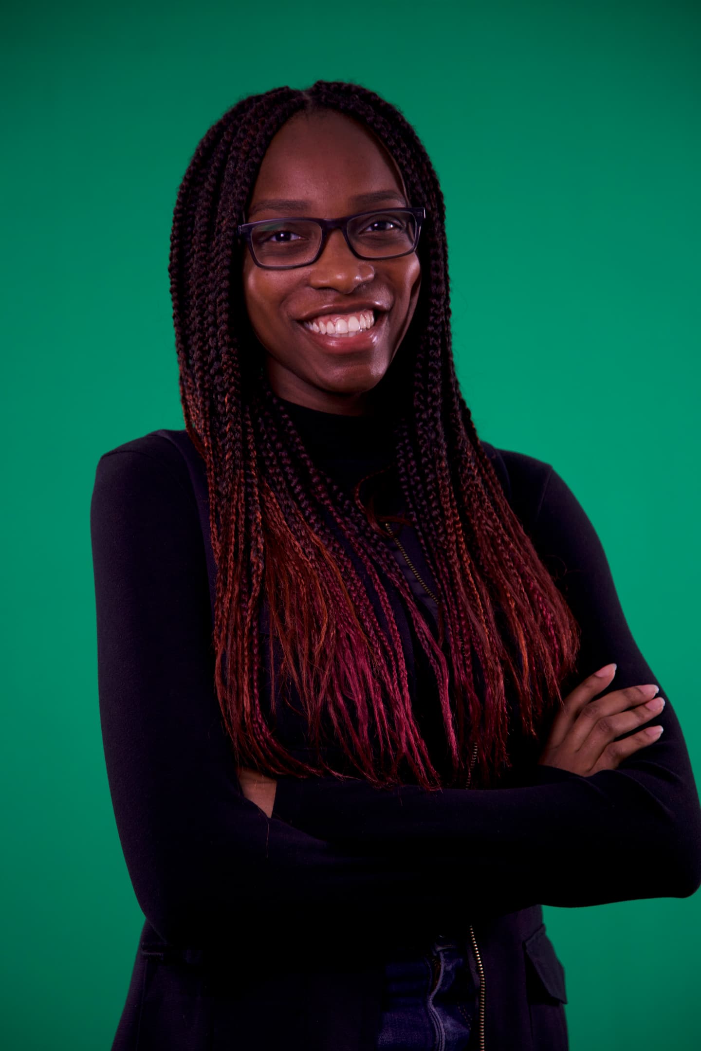 Adeola Sukunbi, a young Black woman with long braids dyed red at the tips, smiles, with her arms crossed, in a black polo neck and glasses, against a green background. 