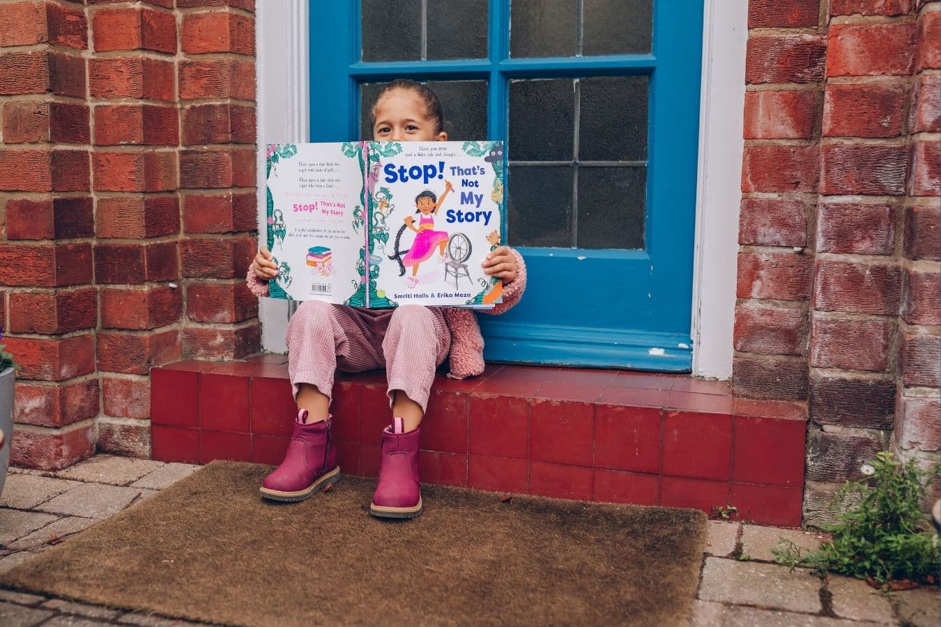 Young mixed-raced child with pink trousers, pink jacket and pink books, sitting on a red doorstep against a blue door. She is hiding the bottom half of her face behind a picture book. 