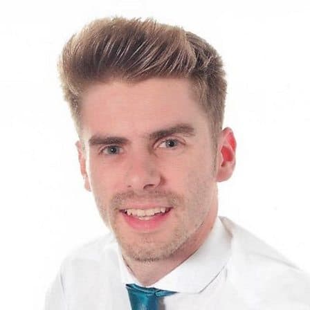 Headshot of a young white man with short blonde hair and stubble. He is wearing a blue tie and a white shirt.