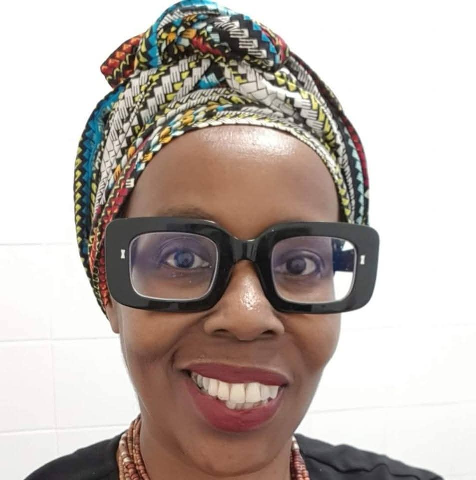 Headshot of a middle-aged Black woman, wearing an African-print headscarf, thick black glasses and red lipstick. She is also wearing a brown beaded necklace and a black top, and is standing against a white background. 