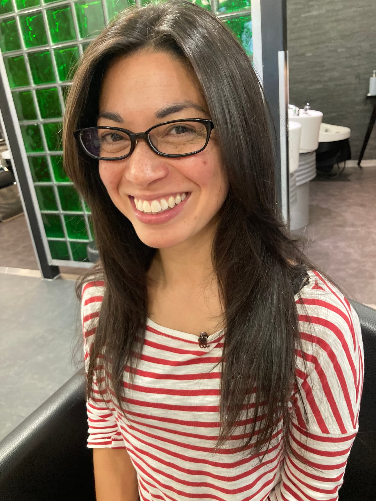 A young, East Asian woman with long, dark-brown hair. She is wearing black glasses and a red and white striped top. She is sitting in a black leather armchair, just outside a bathroom. 