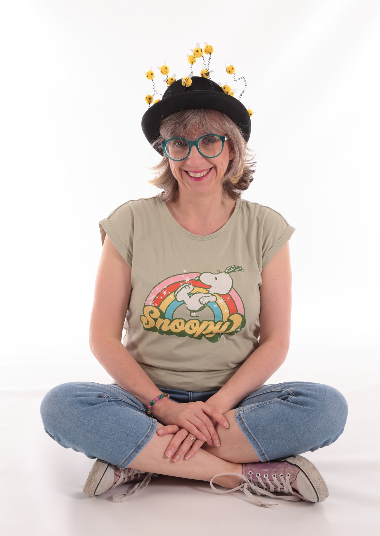 George Kirk, a white woman in her 50s with a grey bob with a fringe and turquoise glasses, sits cross-legged on the floor, smiling broadly. She wears a Snoopy t-shirt, jeans and a black bowler-style hat with whimsical little yellow characters jumping out from it on springs. 