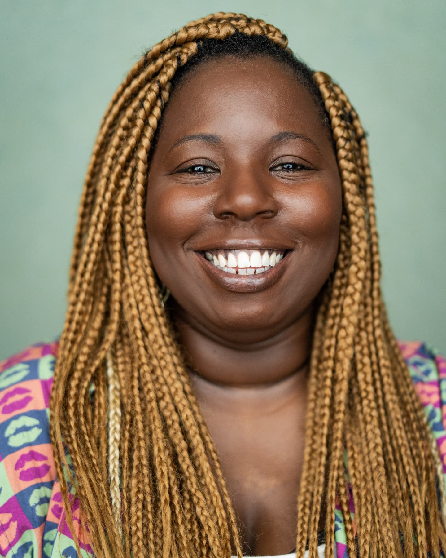 Maame, a thirty-something black woman is pictured in front of a green wall. She is wearing a pink, green and blue shirt patterned with a repeated lip print, over a white vest. She has honey-coloured braids and a wide smile.\n