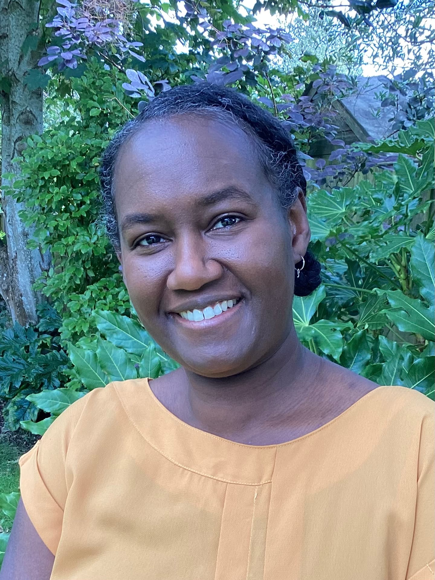 Fabia, a Black woman, is standing outside in front of green plants. She is wearing a yellow top.