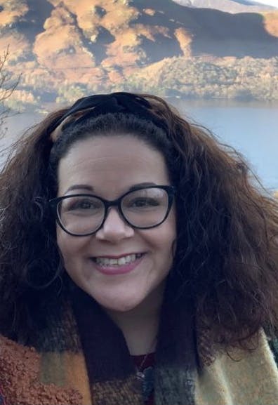 Amy, a forty-something,light-skinned mixed-raced woman, is standing in front of a lake and mountains. She has brown, wavy hair. She is wearing black glasses, a green coat, and an orange and cream scarf.