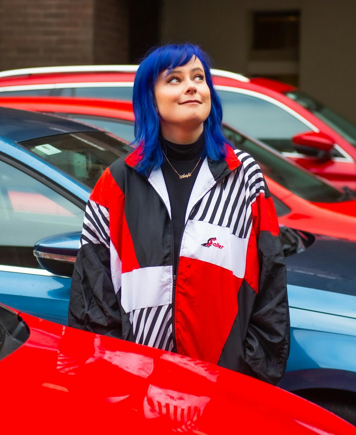 Aoife, a thirysomething white woman stands between a red car and a blue car. She has bright blue hair and is wearing a red, white and black sports jacket.\n\n