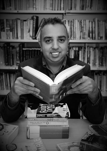 Jake Hope, a man of mixed Asian descent is seen sitting in a school library, surrounding by a range of contemporary books for children and young people.