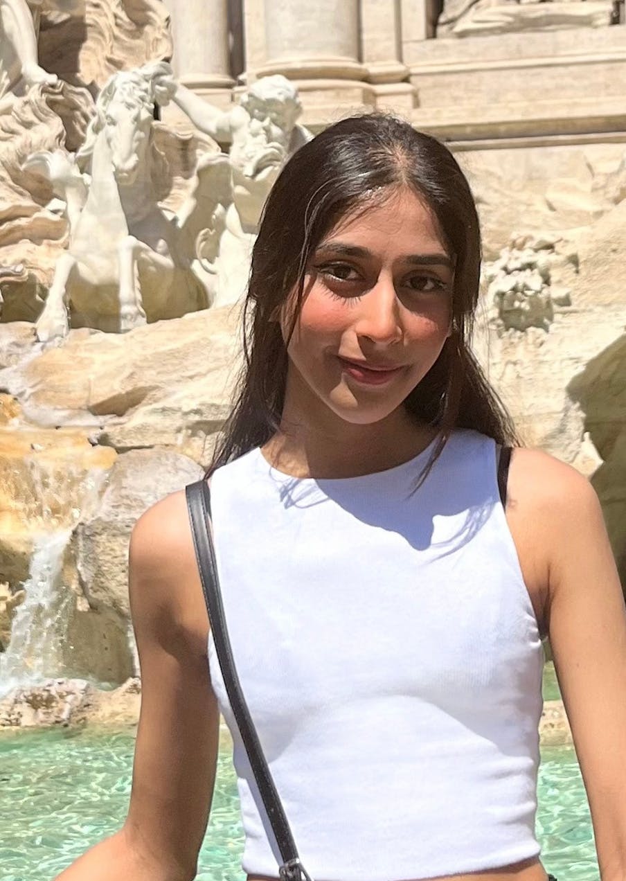 Jayna, a South Asian woman with dark hair, stands in front of a white fountain, wearing a white top with a black crossbody bag.