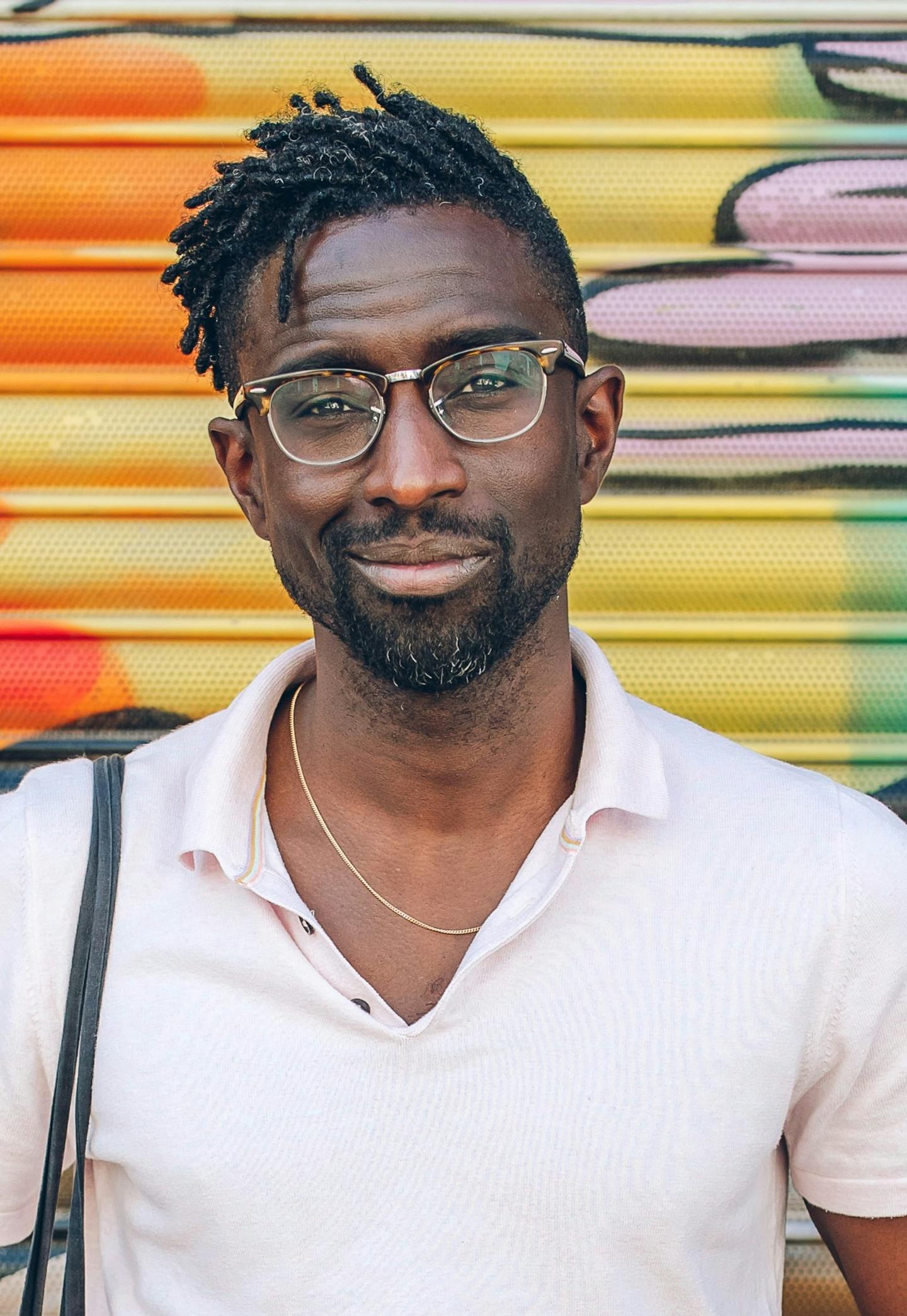 Jeffrey, a fortysomething black man, is standing in front of a a colourful grafittied wall. He is wearing glasses and a pink top. His hair is in locs.