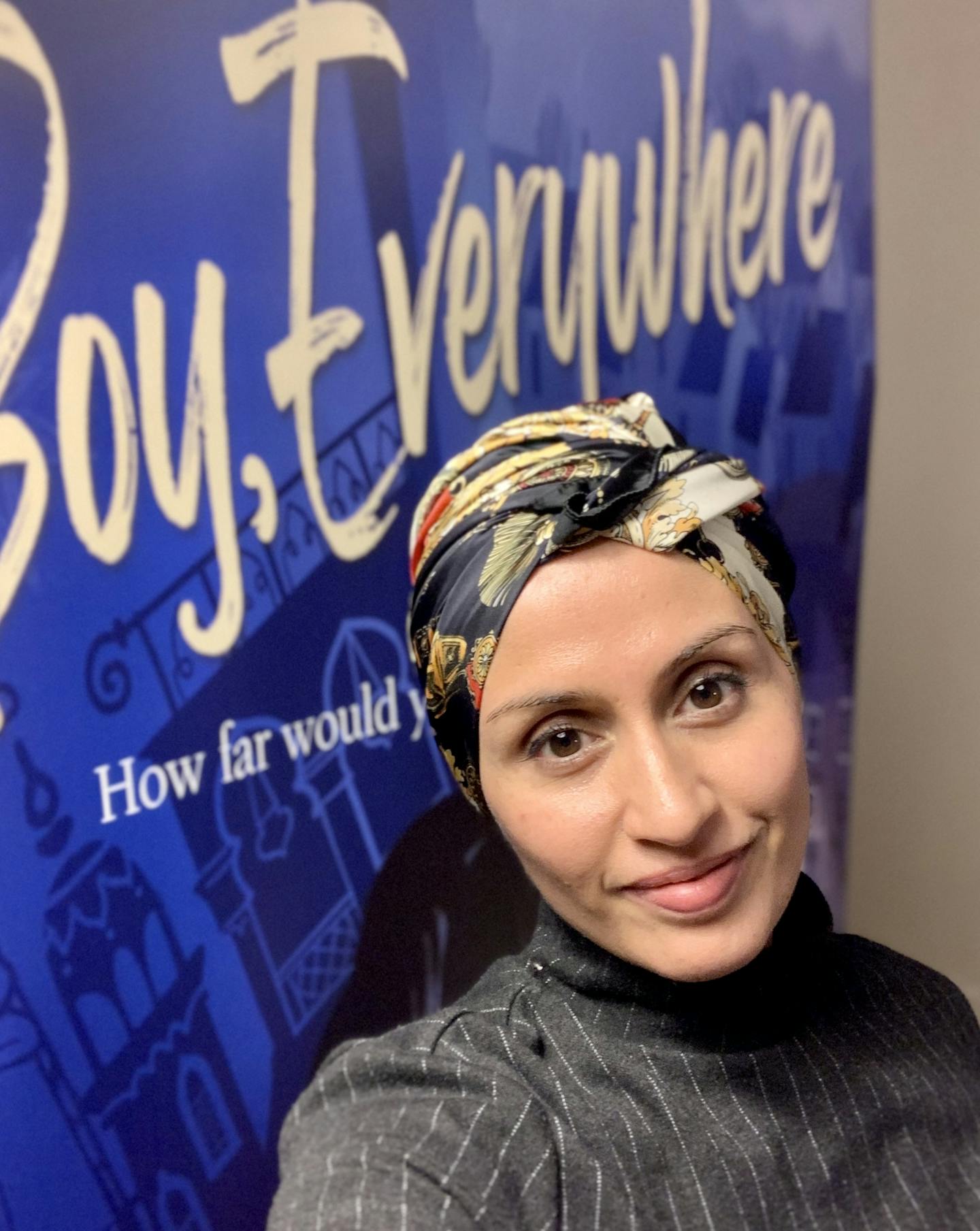 A. M. Dassu, a mixed South Asian woman, is standing in front of a blue banner of her book \nBoy, Everywhere\n, smiling. She is wearing a black pinstripe, high neck top and a black, cream and red patterned satin scarf worn as a head wrap.