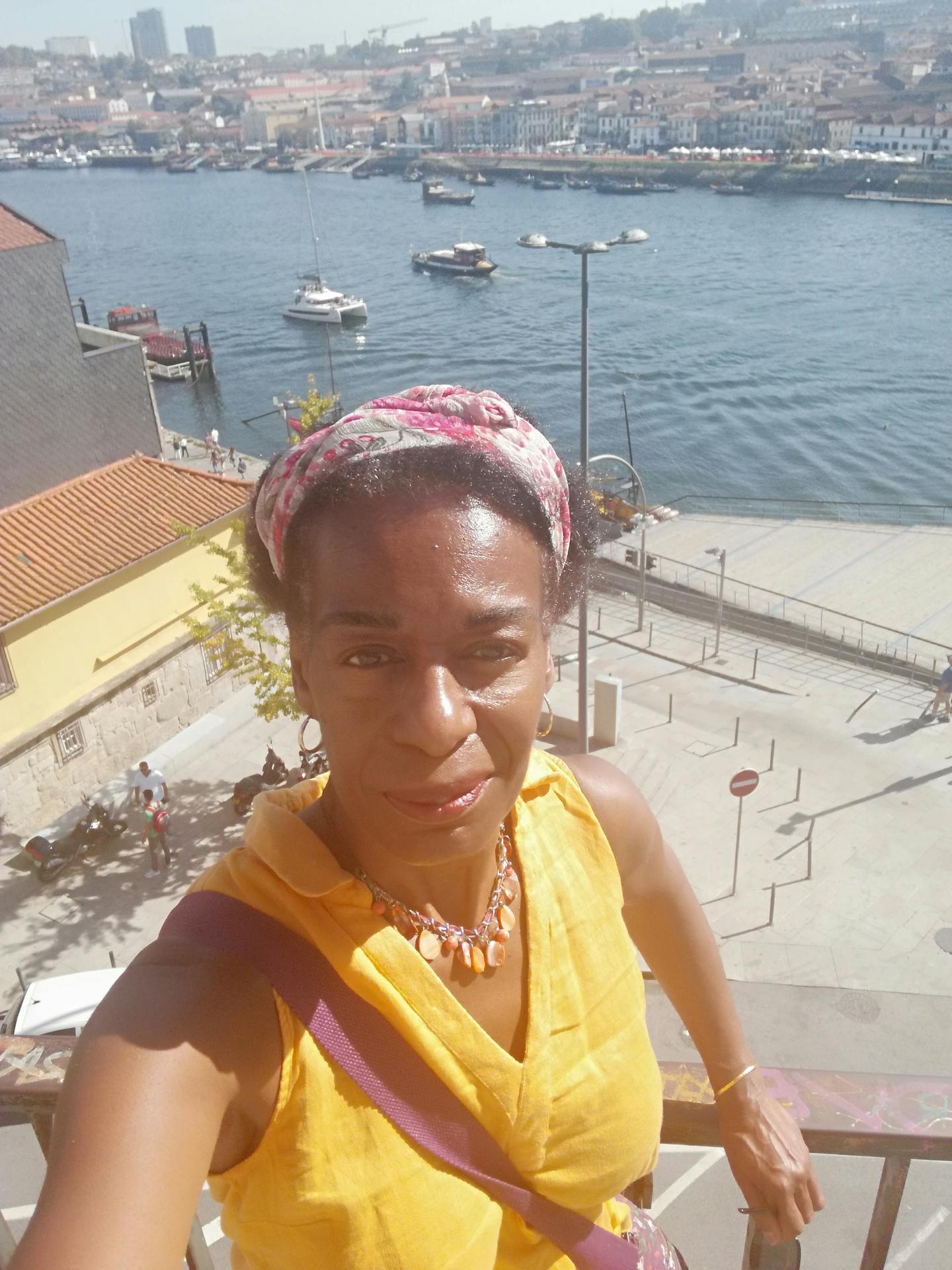Sarah Smith, a Black woman, stands, smiling, in front of a river in Porto, Portugal on a sunny day, wearing a yellow sleeveless blouse and a red colourful bandana covering some of her hair.