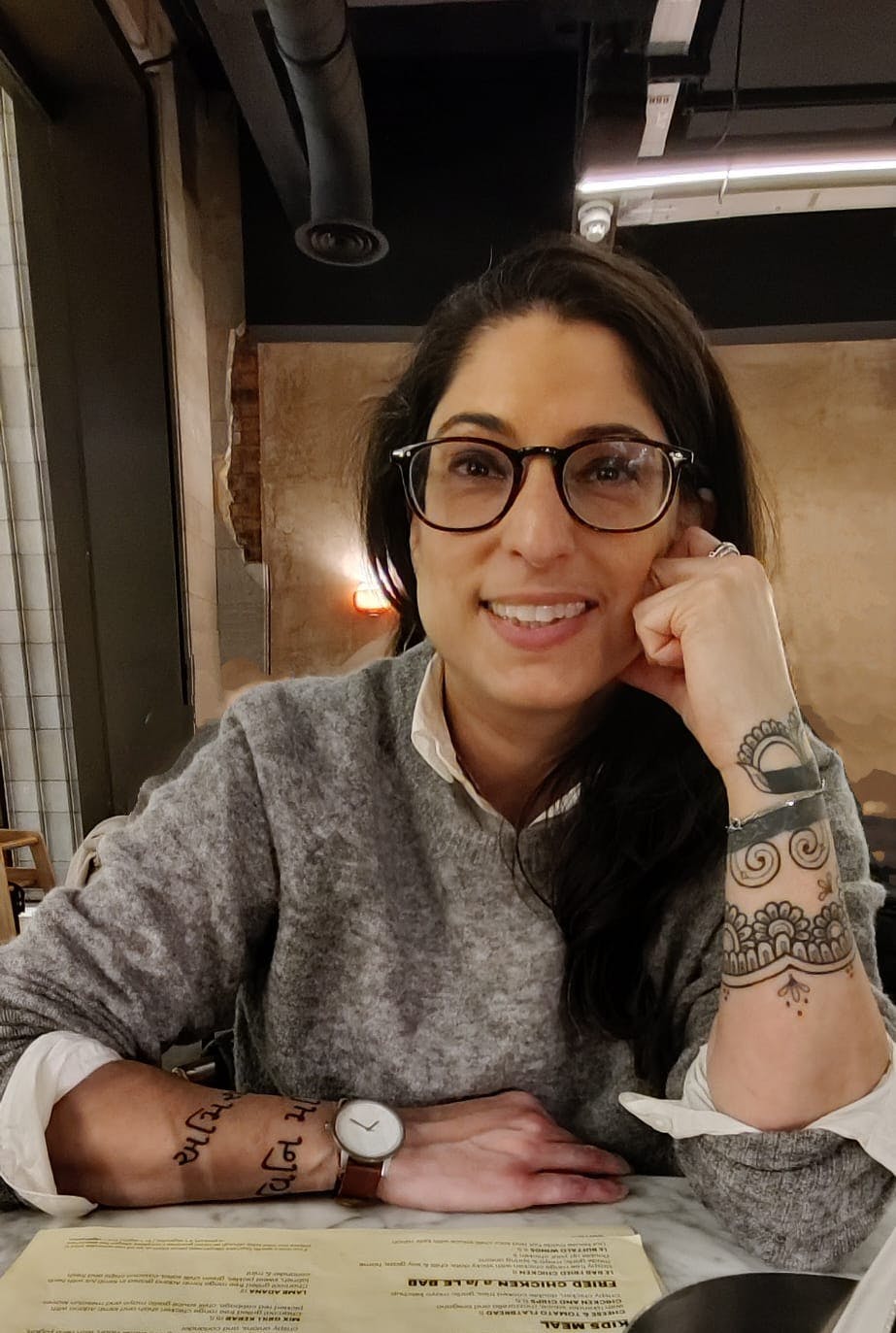 Sinéad, an olive-skinned woman, sits at a table wearing a grey wool jumper. She has dark brown mid length hair, wears glasses and has tattoos on both arms.