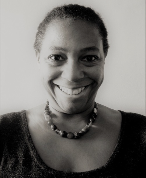 Janet, a middle aged Black woman, is standing in front of a wall in a Black and White, head and shoulders shot. She is smiling and wearing a bead necklace.