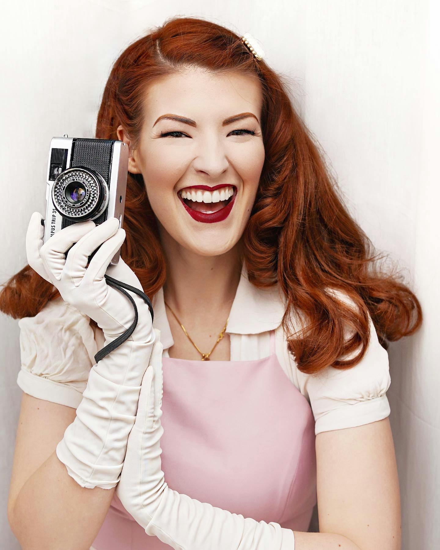 Jessica, a thirtysomething white woman stands in front of a white background. She is wearing a vintage-inspired outfit with gloves and has red hair. She is holding a vintage camera.