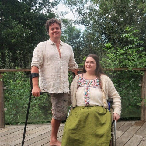 James and Lucy Catchpole are a white couple in their forties, pictured here on some wooden decking with trees behind. She’s a wheelchair-user and he’s an amputee, standing with crutches.