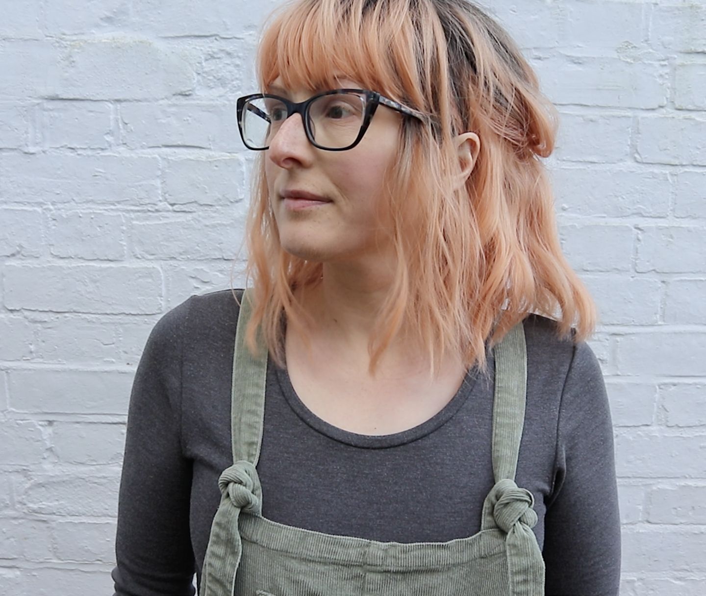 Jen, a thirty something white woman, is standing in front of a white brick wall. She is wearing green dungarees, a grey top, a peach wig and glasses.